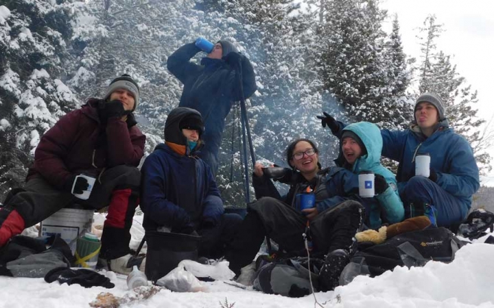 A group of people wearing heavy snow gear smile at the camera while drinking or holding mugs in front of a campfire in the snow.